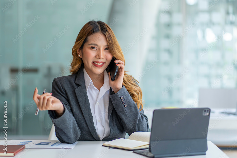Happy business asian manageress working at her desk in the office taking a call on her mobile phone while writing notes on a notepad Happy business asian manageress working at her desk in the office taking a call on her mobile phone while writing notes on a notepad