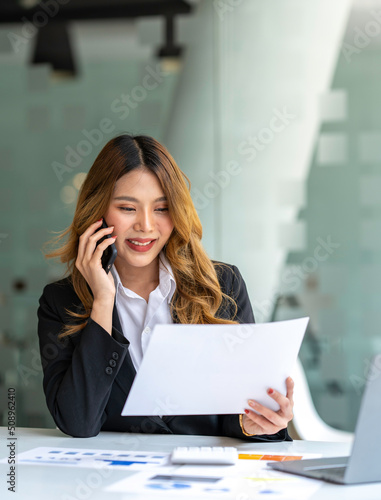 Happy business asian manageress working at her desk in the office taking a call on her mobile phone while writing notes on a notepad