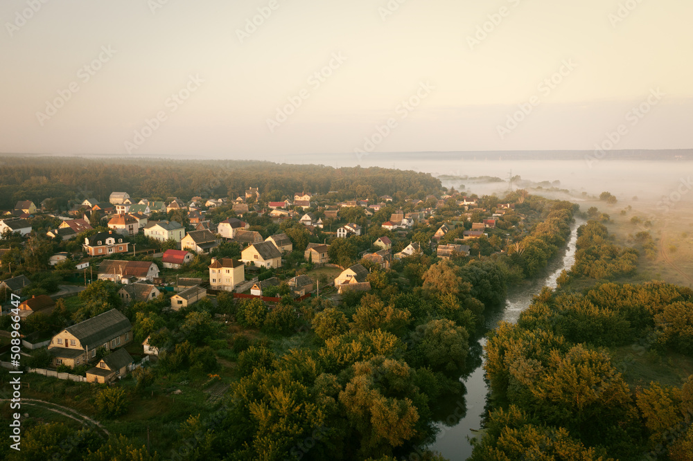 Residential private houses on the banks of the river among the trees on a foggy summer morning - aerial drone shot. Foggy morning in the ecovillage: houses near the water among dense green trees.