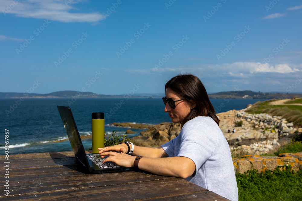 A girl sits on a bench at a wooden table on the ocean, working on a computer. In the background is a thermal mug. In the background is the ocean and mountains.