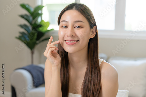 Fresh healthy skin, beautiful smile of asian young woman, girl looking at mirror, applying moisturizer on her face, putting cream treatment before makeup cosmetic routine at home. Facial Beauty. © KMPZZZ