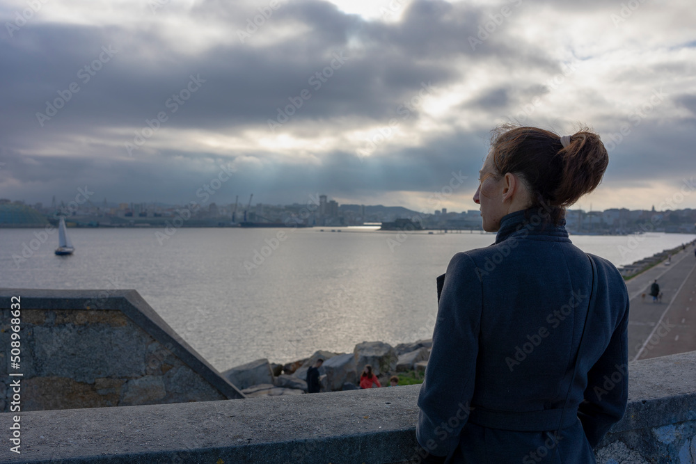 A girl in a gray coat looks at the ocean. Hands in pockets. There is a tail on the head. A sailboat can be seen in the distance. Overcast sky. The clouds are low. Light breaks through the clouds. A girl in a gray coat looks at the ocean. Hands in pockets. There is a tail on the head. A sailboat can be seen in the distance. Overcast sky. The clouds are low. Light breaks through the clouds.