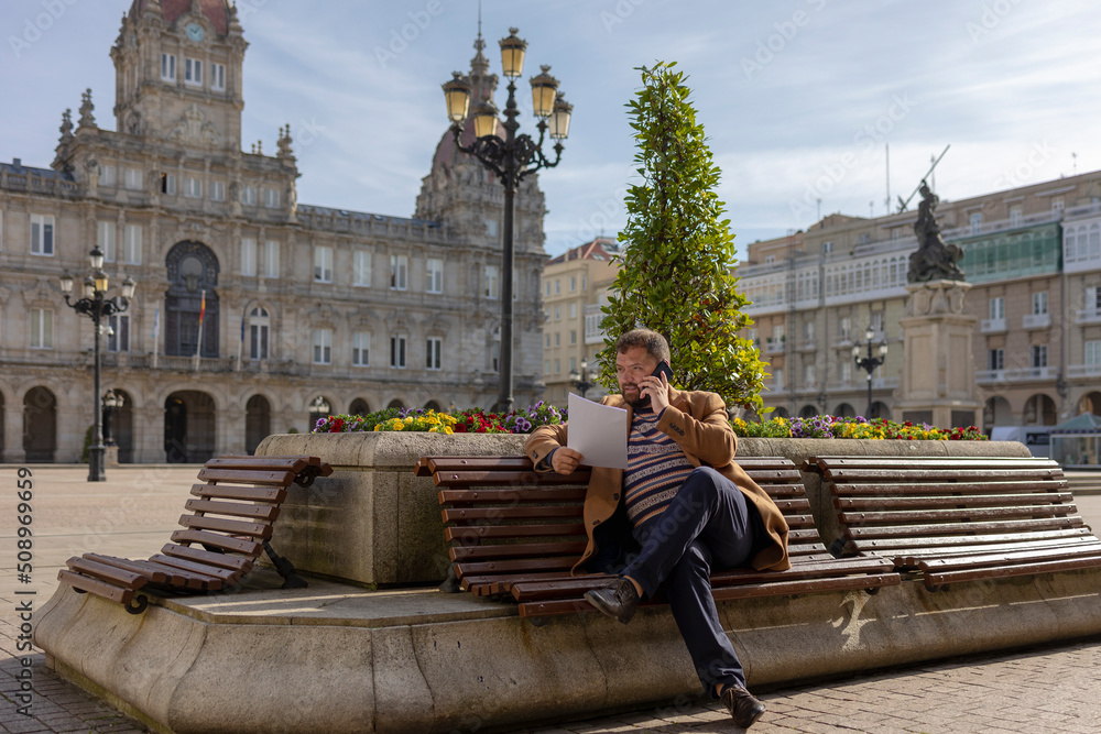 A businessman is talking on the phone, sitting in a park near the city hall. Spain, La Coruna, Melia city, Maria Pita Square. Beautiful facade of the city hall. A man with a beard, wearing a brown