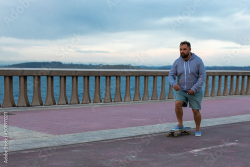 A man with a beard in green shorts, a gray jacket on a skateboard on the embankment. Right foot on the board, left on the ground. The man stares forward intently. Long-range plan. In the background is