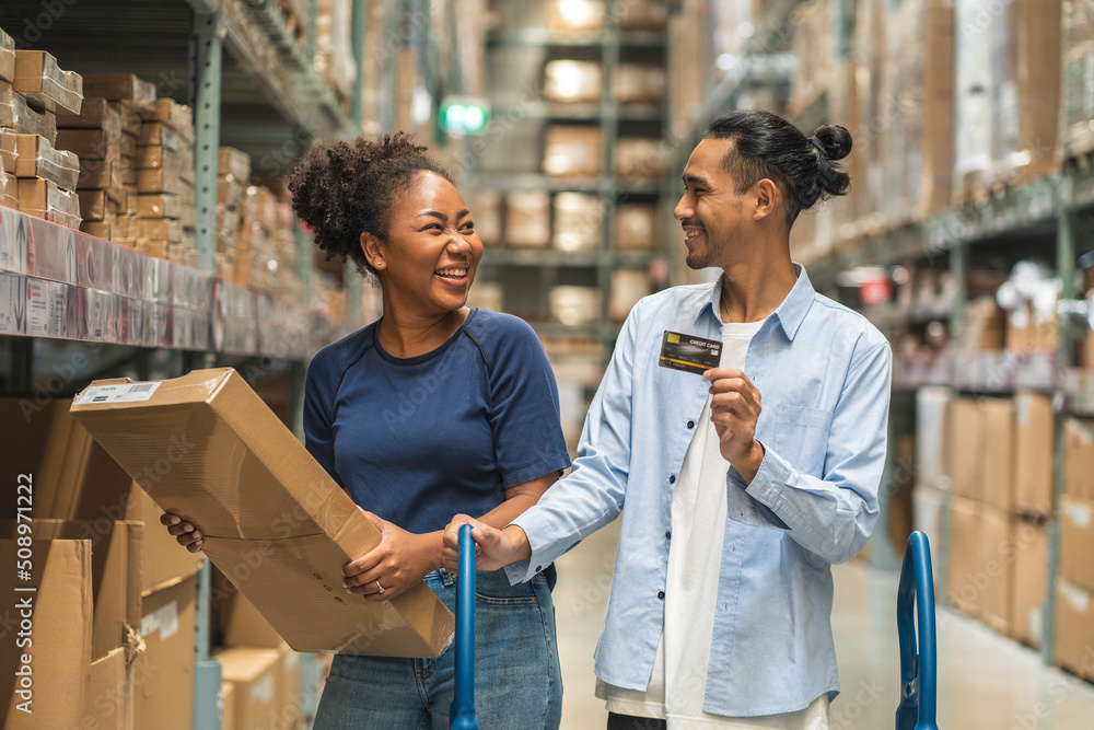 African woman picks up a box of furniture she has bought and an Asian man, her boyfriend holding a credit card, is comfortable to use in a wholesale store warehouse.