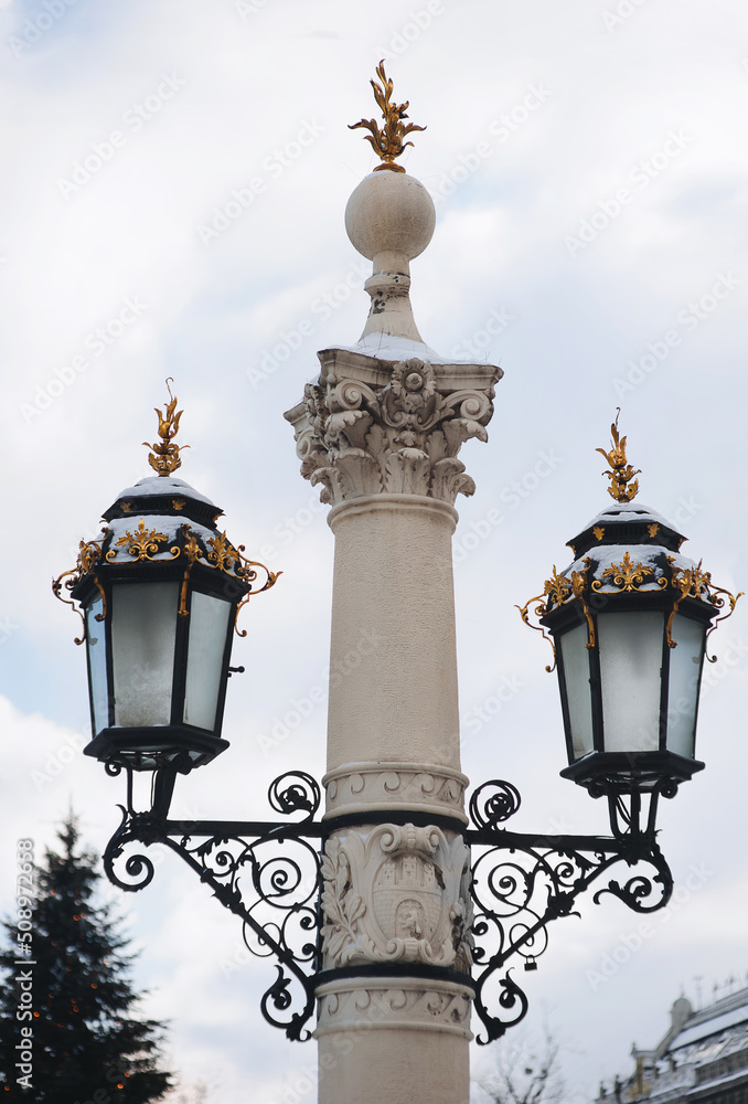 Two old large black lanterns on a thick column with a capital and gilded tops in the historical center of Lviv, near the Opera and Ballet Theatre.