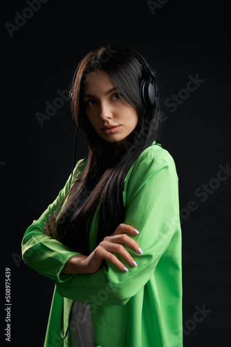 Attractive brunette lady looking at camera with earphones on head indoors. Front view of calm girl in stylish jacket with crossed hands, listening music, isolated on black. Concept of lifestyle. 