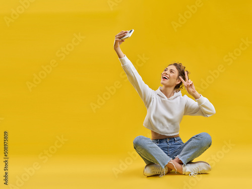 Foto Young girl taking selfie with smartphone, sitting with crossed legs in studio