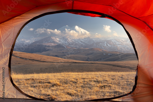 View from open door of red tent to snow-covered Mount Elbrus at sunset golden light, Caucasus Russia