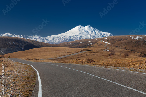 Winding road leads through hills to snowy mountains against clear blue sky, Caucasus, Russia