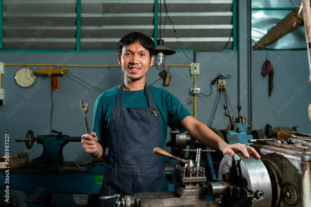 A craftsman is using a small tool to tap the iron that is still attached to the lathe, which he had machined before, whether the quality has been reached, in his small workshop. A craftsman is using a small tool to tap the iron that is still attached to the lathe, which he had machined before, whether the quality has been reached, in his small workshop.