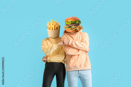 Couple with tasty french fries and burger instead of their heads bumping fists on light blue background photo