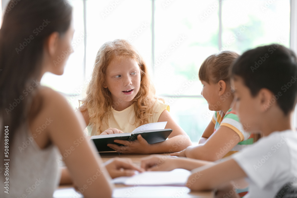 Fototapeta premium Little redhead girl reading book during literature lesson in classroom