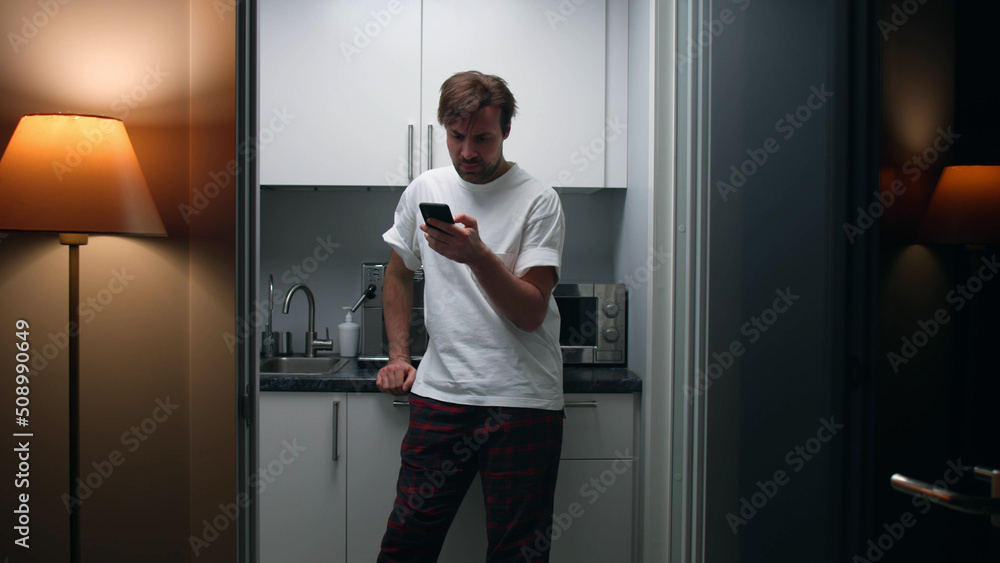 Tired guy chat on smartphone standing in kitchen late in evening Tired guy chat on smartphone standing in kitchen late in evening