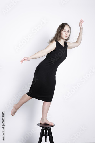 girl in a black dress and with natural make-up keeps balance while standing on a black chair