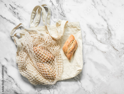 Reusable eco bags and buns on a light marble background, top view