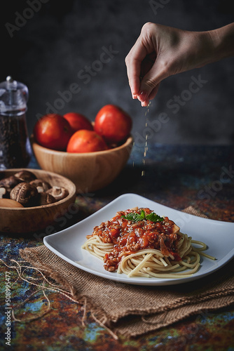 Female chef hand seasoning a plate of juicy spaghetti with tomato sauce on dark background.
