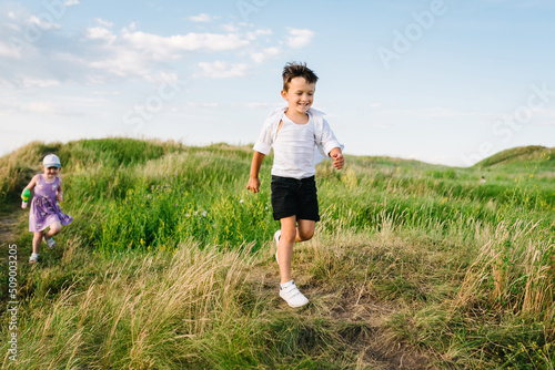 The happy little children running and playing in a field and mountains on a sunny summer day at sunset. Cute kids laugh and walk on green grass in the spring.