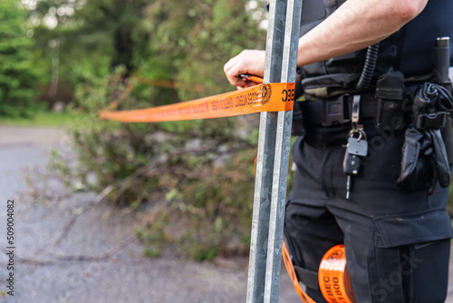 Closeup selective focus view on belt of local police officer closing a main road with tape in aftermath of storm, blurry uprooted tree in background.
