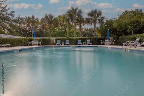 Swimming pool with lounge chairs, palm trees, and hedge. Long exposure creates a smooth effect on the water's surface.