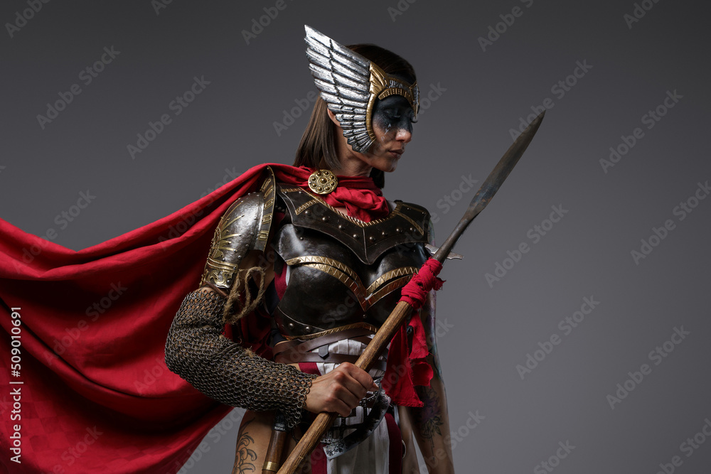 Obraz premium Shot of female barbarian holding spear dressed in steel armor with helmet against grey background.