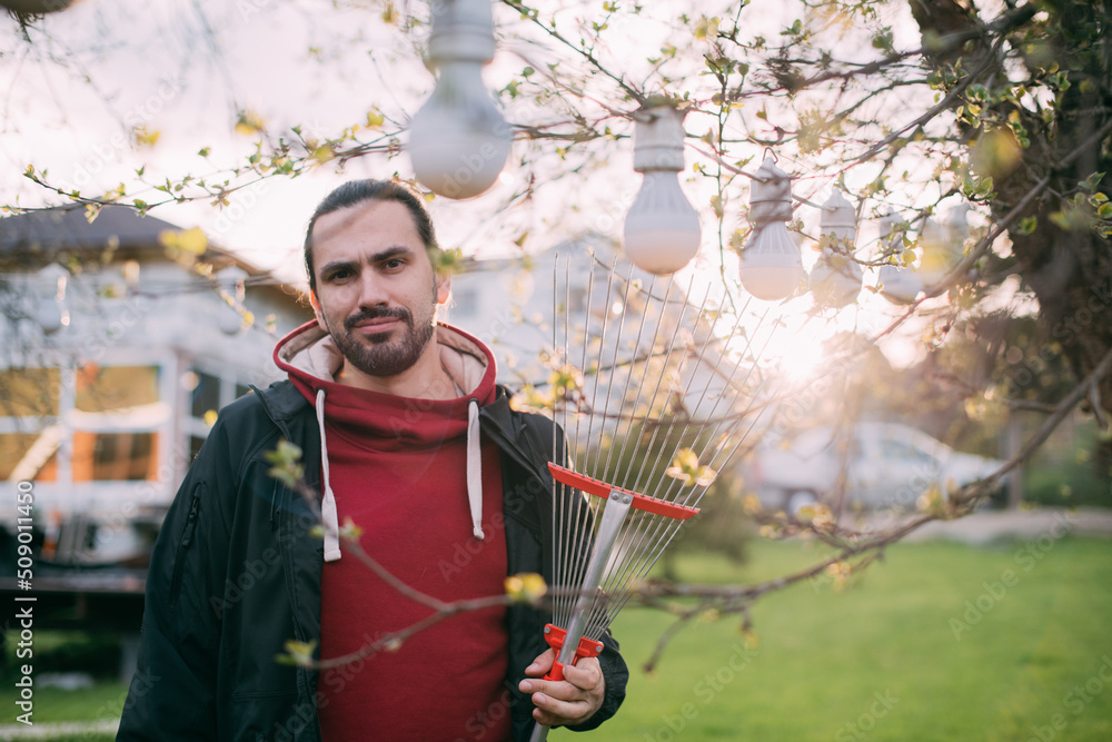 Portrait of a gardener - a young man with a fan rake in his hands in the garden in the evening at sunset.