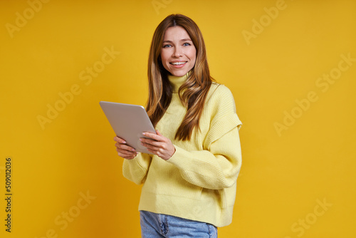 Happy young woman holding digital tablet while standing against yellow background