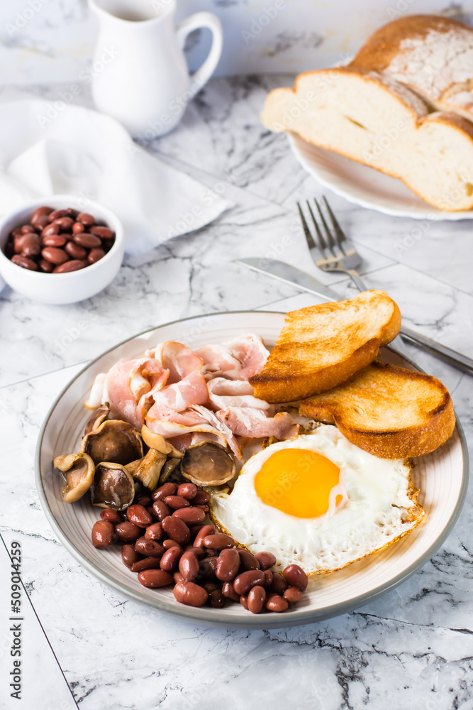 English breakfast with fried egg, bacon, beans, mushrooms and toast on a plate. Vertical view English breakfast with fried egg, bacon, beans, mushrooms and toast on a plate. Vertical view