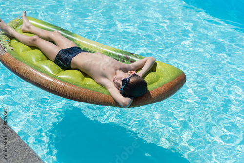 Boy sunbathing squinting his eyes from the sun and lying on a mattress in a pool of blue water