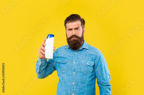 brutal bearded man hold shampoo bottle on yellow background, haircare