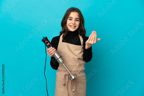Little girl using hand blender isolated on blue background inviting to come with hand. Happy that you came