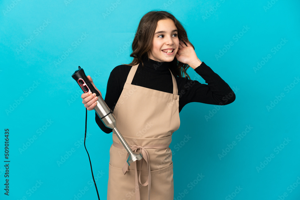 Little girl using hand blender isolated on blue background listening to something by putting hand on the ear Little girl using hand blender isolated on blue background listening to something by putting hand on the ear