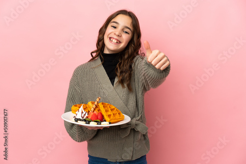 Little girl holding waffles isolated on pink background with thumbs up because something good has happened