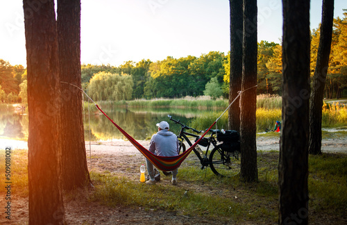 Man travels on bicycle, relaxing in red hammock, rest in forest near lake. Cyclist in hammock at campsite by river. Male on bike in hammock at sunset.