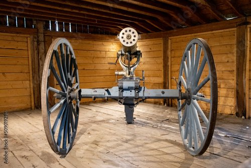 old military gatling gun from the settlement era on wooden cart inside illuminated shed photo