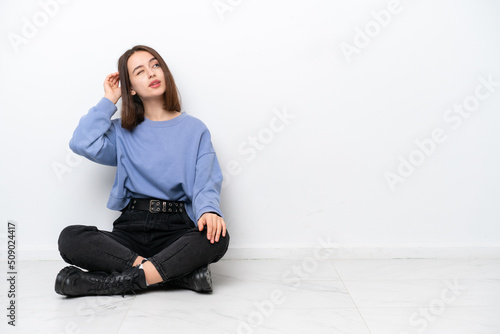 Young Ukrainian woman sitting on the floor isolated on white background having doubts and with confuse face expression