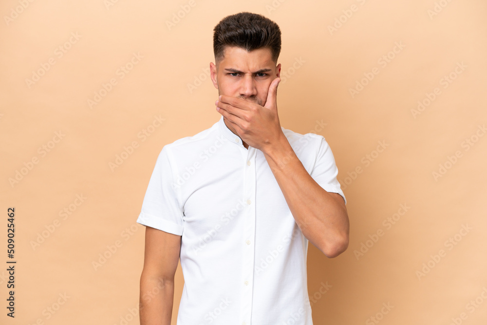 Young caucasian man isolated on beige background covering mouth with hand Young caucasian man isolated on beige background covering mouth with hand