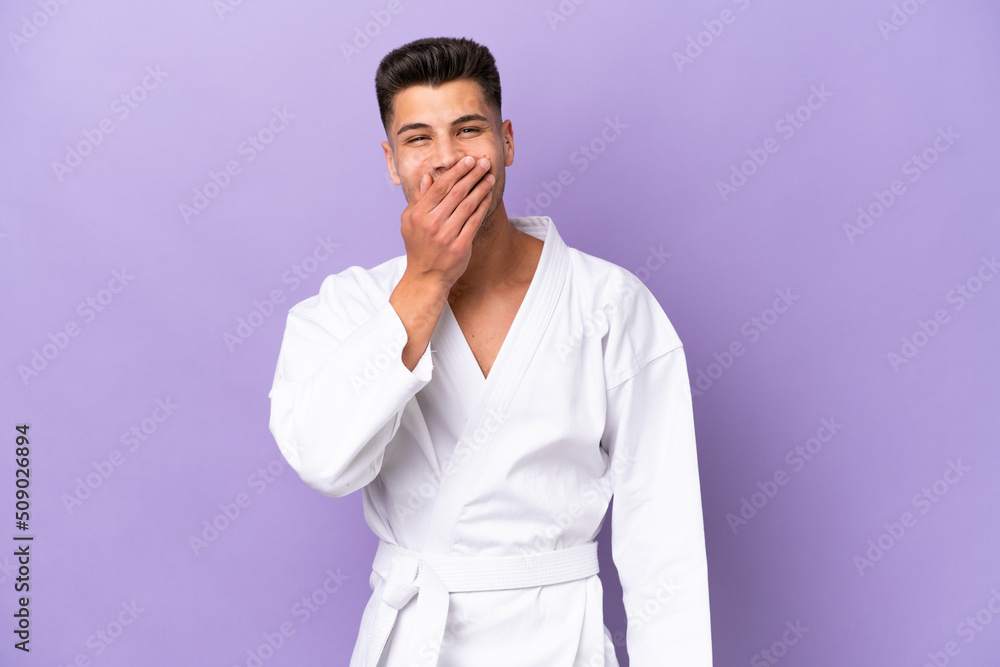 Young caucasian man doing karate isolated on purple background happy and smiling covering mouth with hand Young caucasian man doing karate isolated on purple background happy and smiling covering mouth with hand