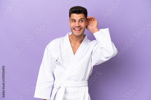 Young caucasian man doing karate isolated on purple background listening to something by putting hand on the ear