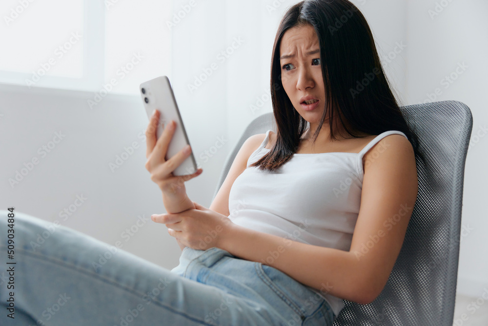 Irritated shocked young Asian woman read fake bad news in messages sit on chair at home interior living room. Distance communication, Remote work, Social media concept. Cool offer Banner Mockup Irritated shocked young Asian woman read fake bad news in messages sit on chair at home interior living room. Distance communication, Remote work, Social media concept. Cool offer Banner Mockup