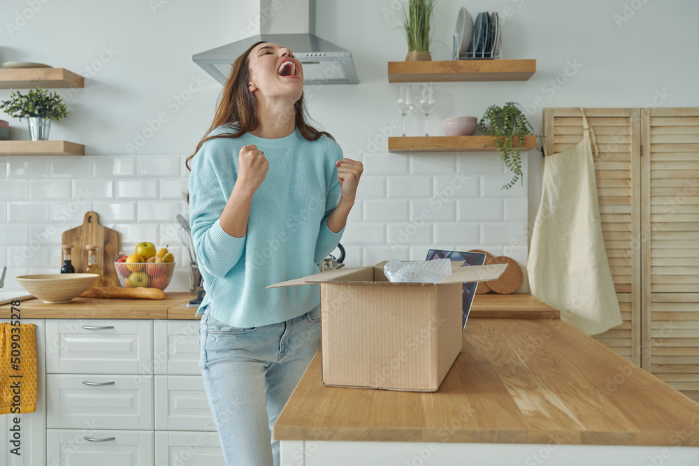 Excited young woman unpacking box and gesturing while standing at the domestic kitchen Excited young woman unpacking box and gesturing while standing at the domestic kitchen
