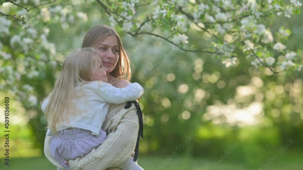 A young cheerful mum is holding a baby in her arms in a beautiful blooming garden while on a summer holiday. Family outdoor lifestyle. A young cheerful mum is holding a baby in her arms in a beautiful blooming garden while on a summer holiday. Family outdoor lifestyle.