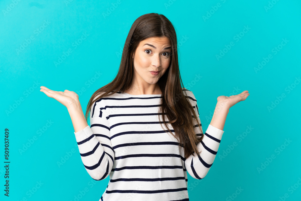 Teenager Brazilian girl over isolated blue background having doubts while raising hands Teenager Brazilian girl over isolated blue background having doubts while raising hands