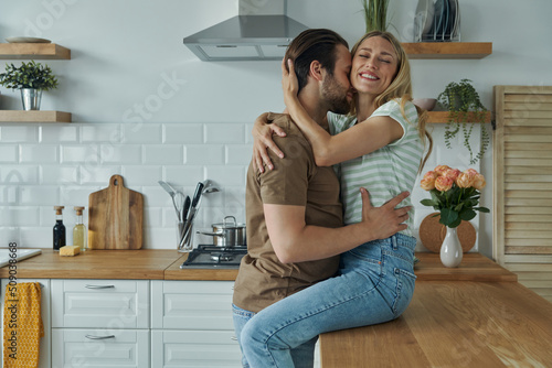 Beautiful young couple embracing and kissing while woman sitting on the kitchen counter