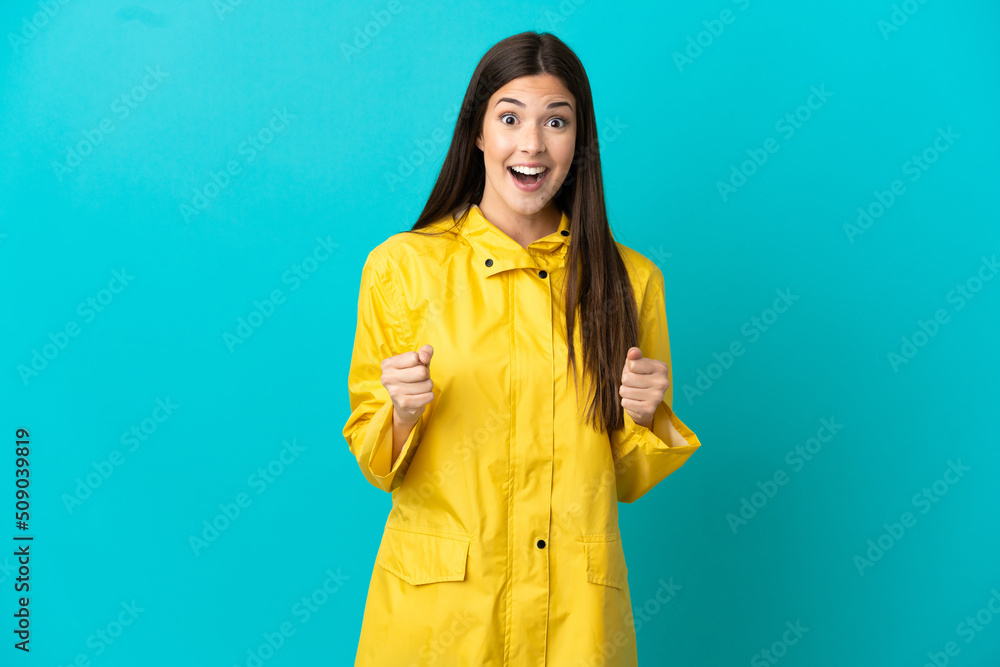 Teenager Brazilian girl wearing a rainproof coat over isolated blue background celebrating a victory in winner position Teenager Brazilian girl wearing a rainproof coat over isolated blue background celebrating a victory in winner position