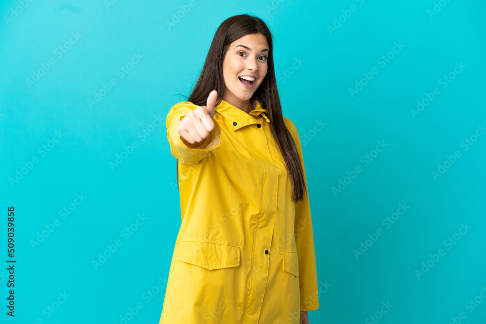 Teenager Brazilian girl wearing a rainproof coat over isolated blue background with thumbs up because something good has happened Teenager Brazilian girl wearing a rainproof coat over isolated blue background with thumbs up because something good has happened