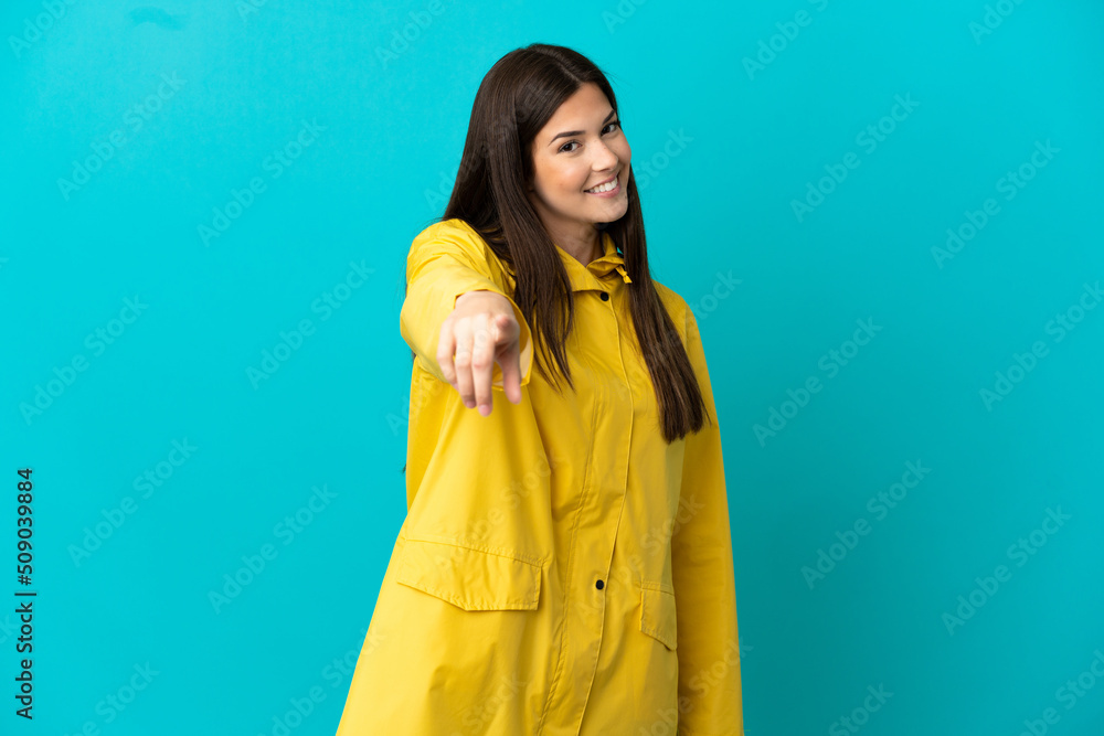 Teenager Brazilian girl wearing a rainproof coat over isolated blue background points finger at you with a confident expression