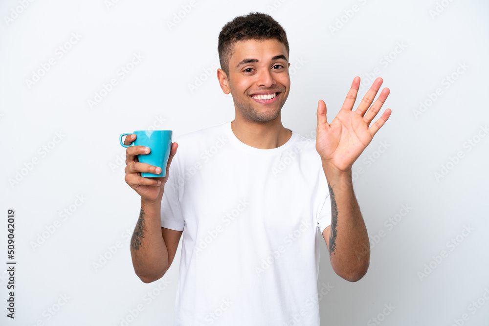 Young Brazilian man holding cup of coffee isolated on white background saluting with hand with happy expression Young Brazilian man holding cup of coffee isolated on white background saluting with hand with happy expression