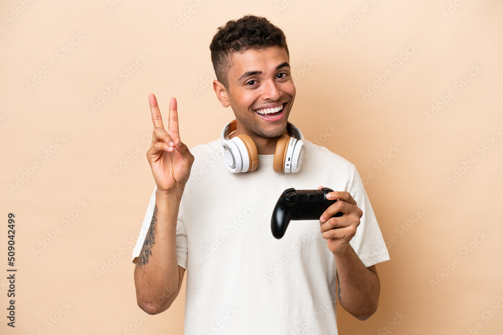 Young Brazilian man playing with a video game controller isolated on beige background smiling and showing victory sign Young Brazilian man playing with a video game controller isolated on beige background smiling and showing victory sign