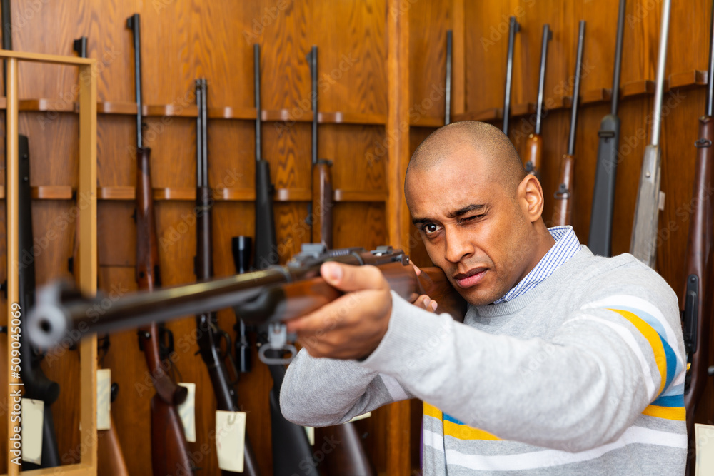 Focused latin american man testing hunting shotgun in armory shop, imitating aiming
